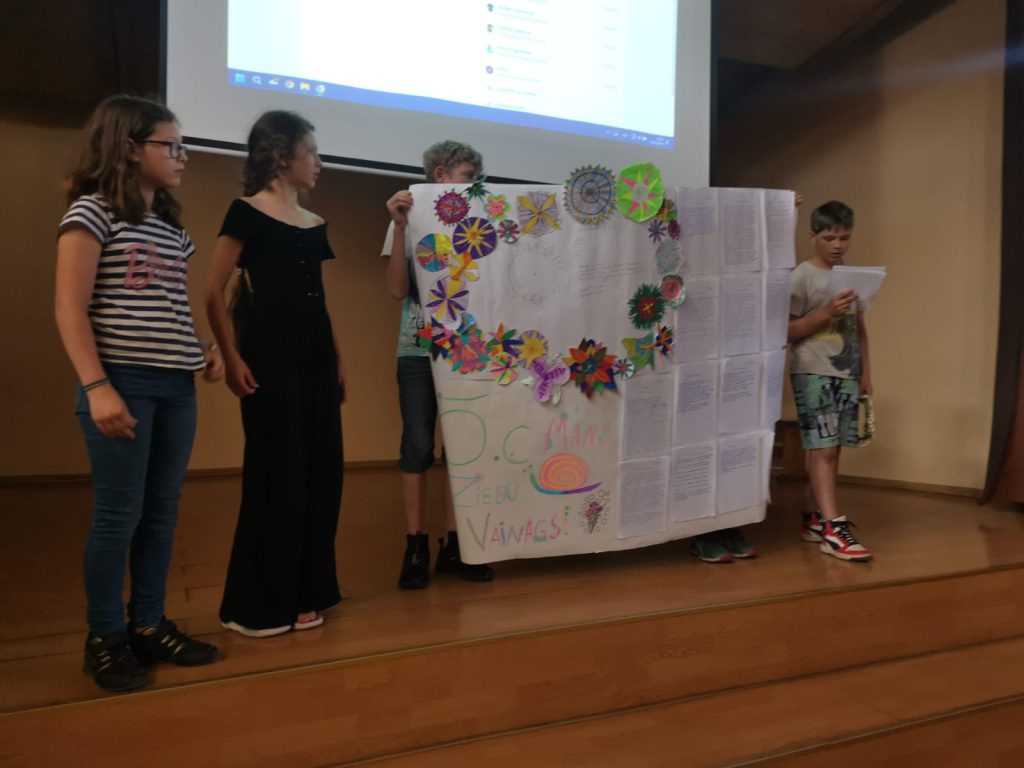 Skolēni prezentē, plakāts, ziedu vaonags, projektors, skatuve, zēns lasa no lapas
