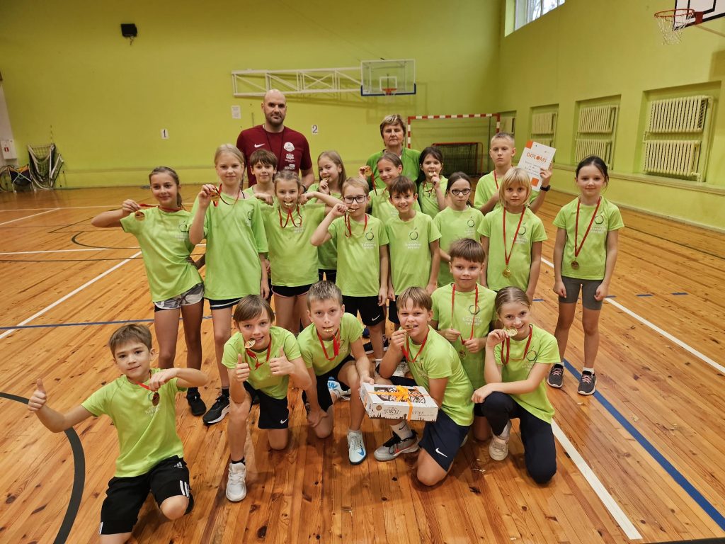 Skolēni un skolotājs sporta zālē, zaļos krekliņos priecīgi pozē